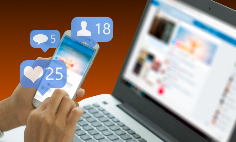 A person holding a smartphone showing social media notification icons, with a laptop in the background, representing Social Media Graphic Design Services.